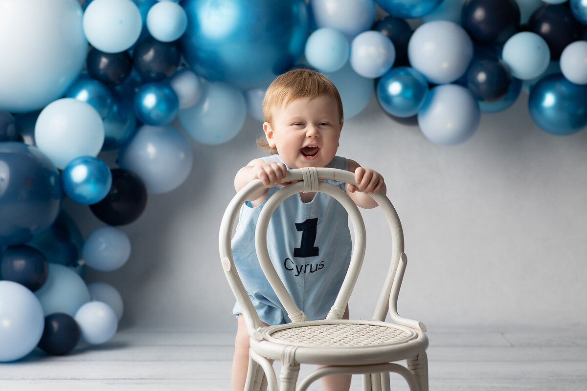 A joyful baby boy in a personalized romper with the number one stands behind a white chair, beaming with excitement in front of a festive blue balloon arch. Captured by a Dallas baby milestone photographer.