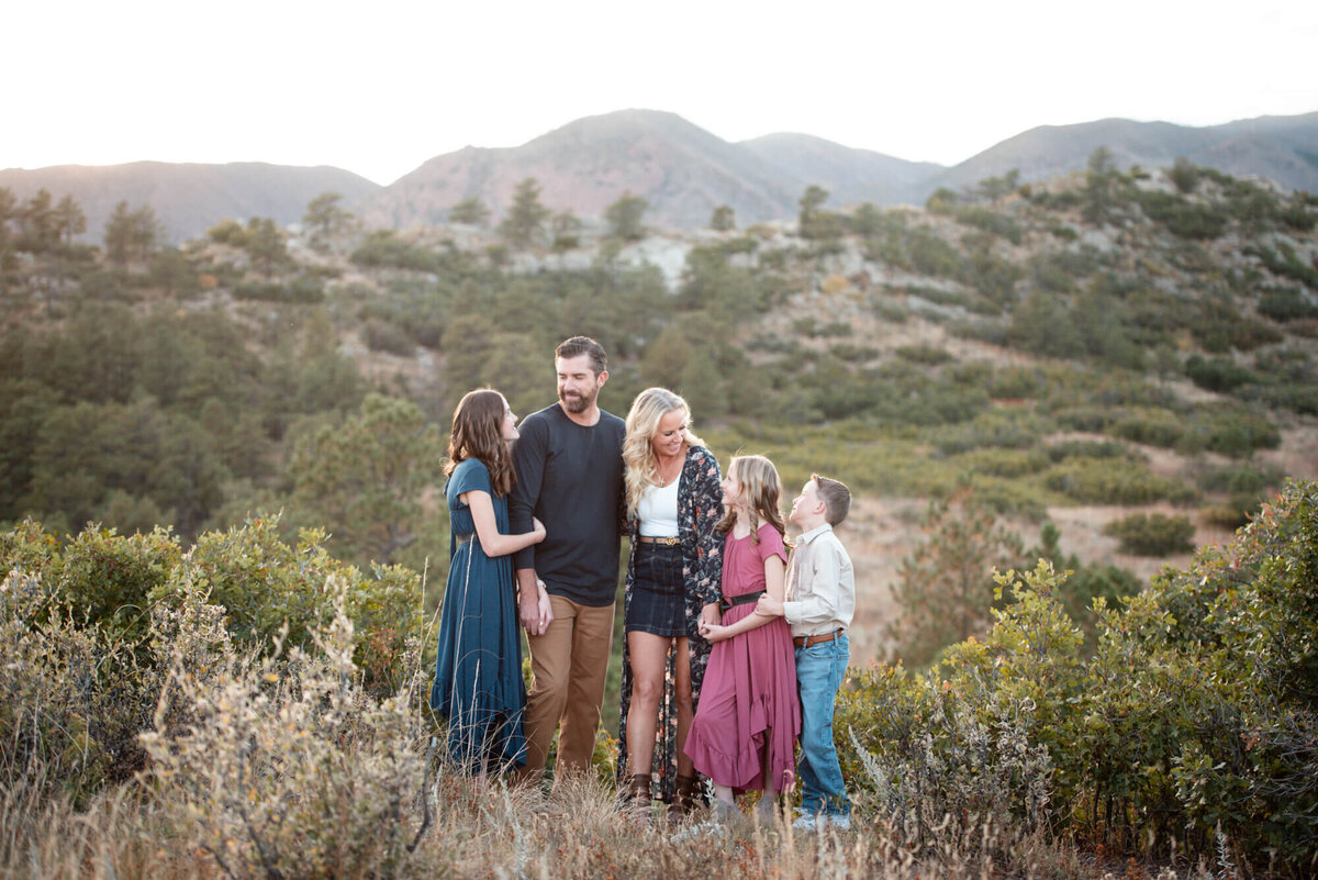 A family of 5 laughs together while exploring a mountain trail at sunset and hugging each other