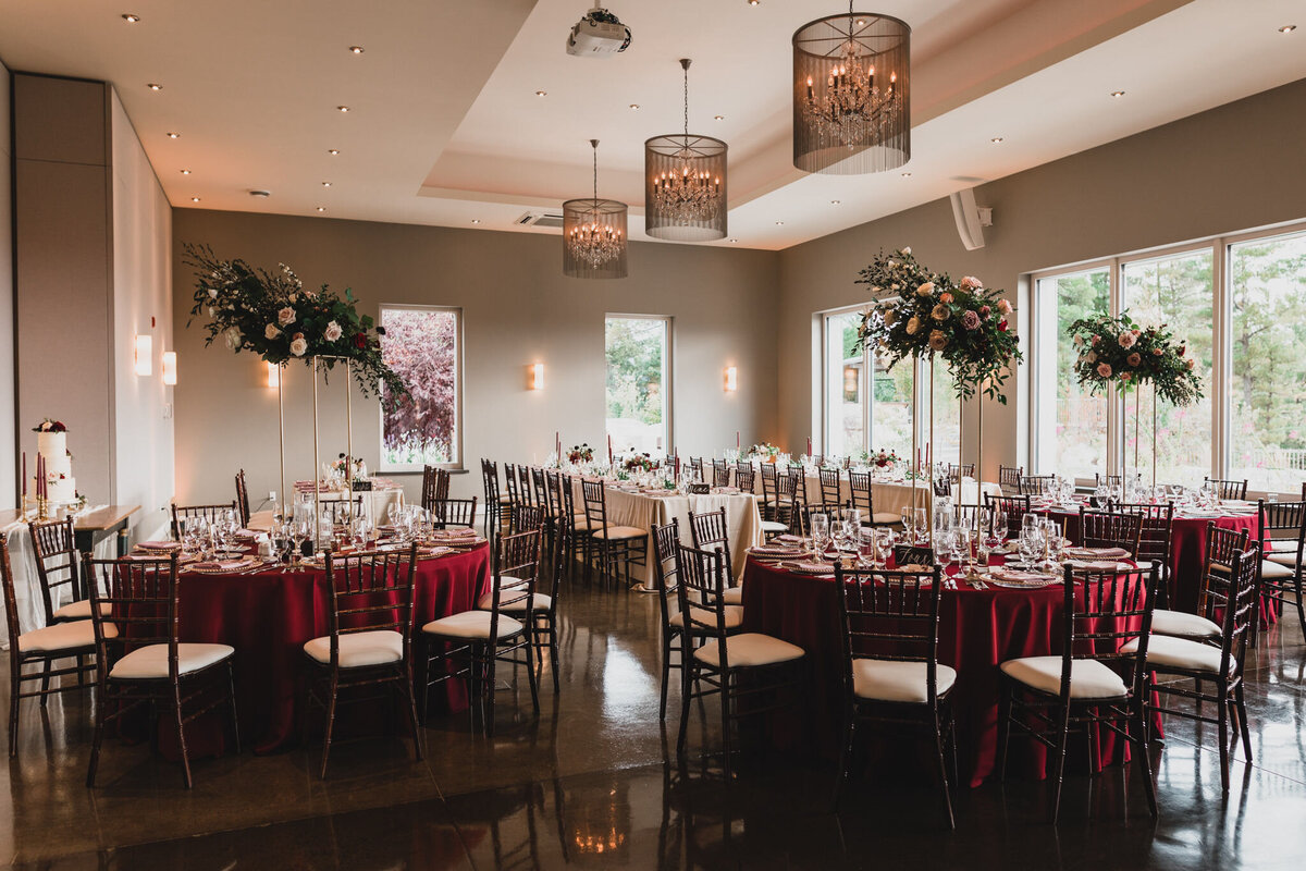 Dark burgundy linens with black metal dining chairs and striking tall floral centrepieces are inside the wedding venue Le Belvedere in Wakefield Quebec