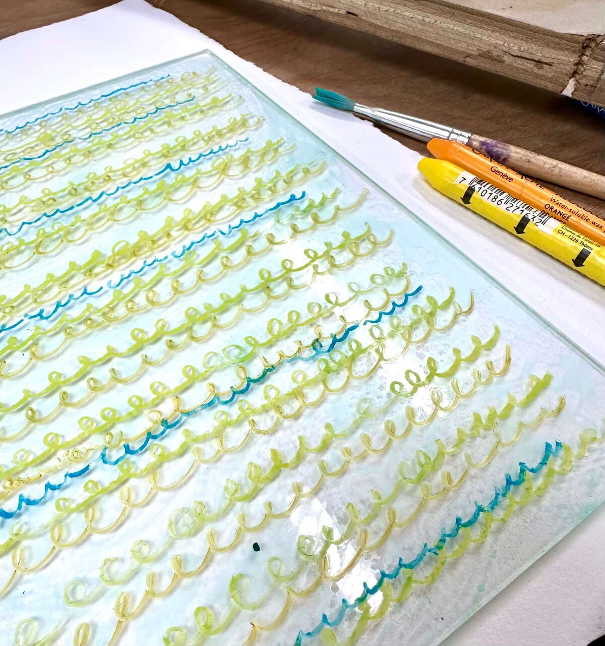 A detailed shot of layered mark-making on a plexiglass plate using yellow, green, and blue wax crayons, ready for print.