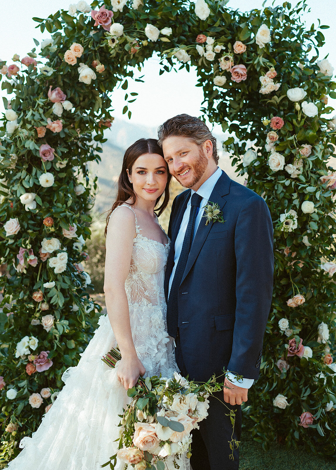 bride and groom standing in front of an arch that is filled with lush floral and greenery