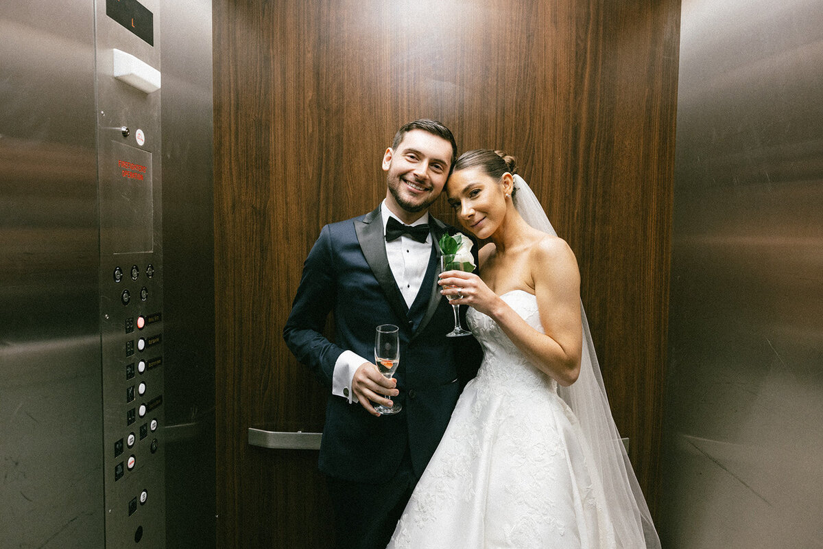 Bride and groom pose in the elevator, champagne glasses in hand after their wedding ceremony. 
