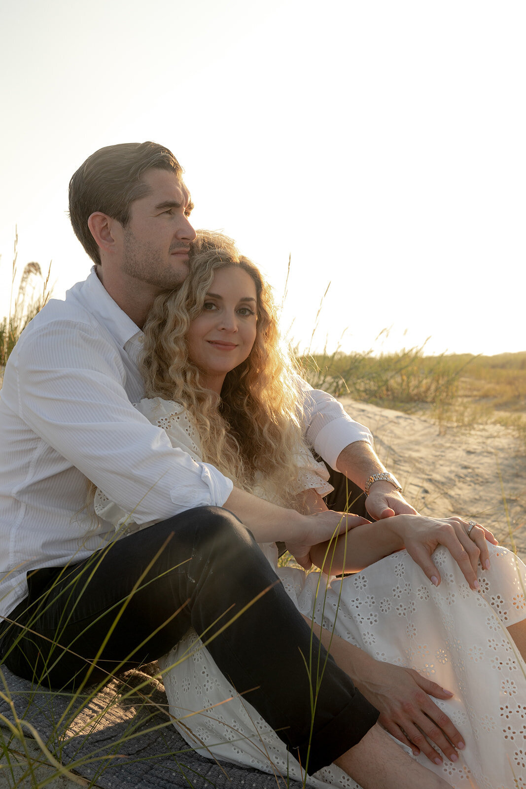 Couple sitting at Charleston beach. Woman  is leaning into man while sitting between his legs. She smiles into the camera. The man is looking straight . Both are surrounded by sand and grass.