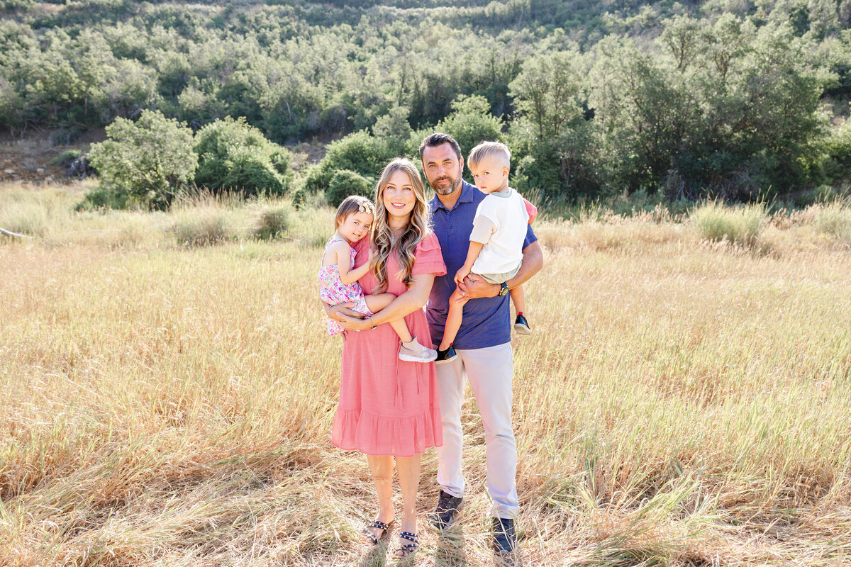 A family of four poses together in an open field, with rolling hills and trees in the background. The parents hold their two young children close, their outfits blending beautifully with the natural scenery.