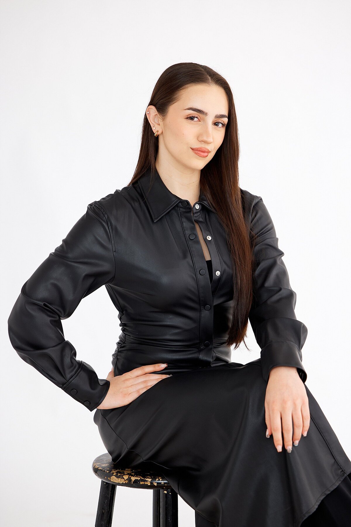 ophisticated young woman with sleek, straight hair wearing a full-length black faux leather dress, posing confidently on a stool with a white backdrop for real estate


