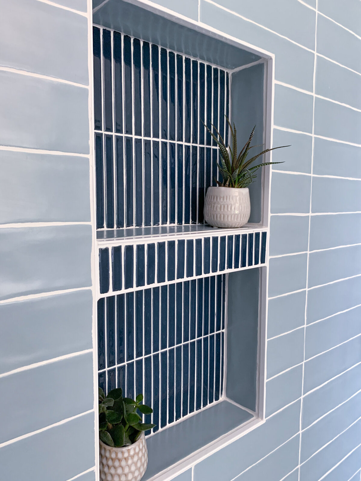 A close-up of a built-in shower niche tiled with dark blue vertical tiles and white grout. The niche has two shelves, also outlined in white tile. The surrounding shower walls are tiled with horizontal rows of light blue rectangular tiles and white grout. On the top shelf, a small white pot with a textured surface holds a succulent plant with spiky green leaves. On the bottom shelf, a small gray and white patterned pot contains a lush green leafy plant. The lighting is bright and even, highlighting the tile work and the plants.
