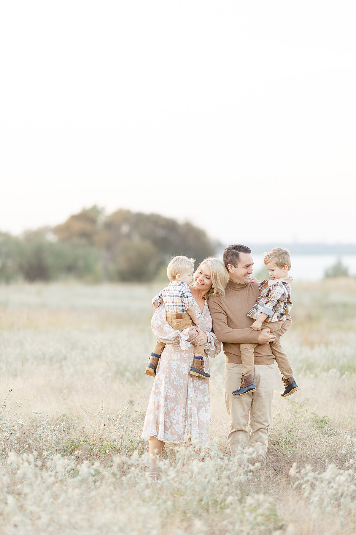 Beautiful family portrait of a mother and father holding their two young boys as they pose for family photos in the middle of an open field at a Dallas park.