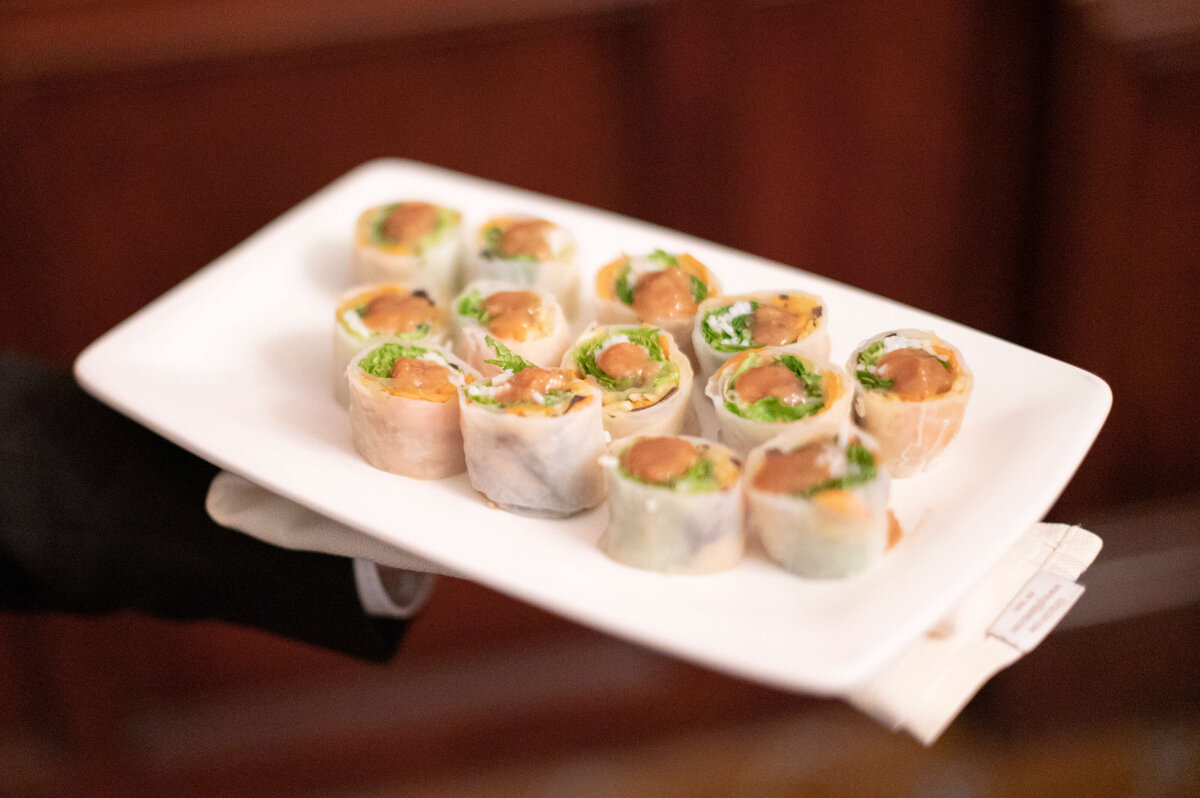 A person holds a rectangular white plate with neatly arranged sushi rolls. The rolls are filled with greens and a brown topping, presented elegantly. The background is softly blurred.