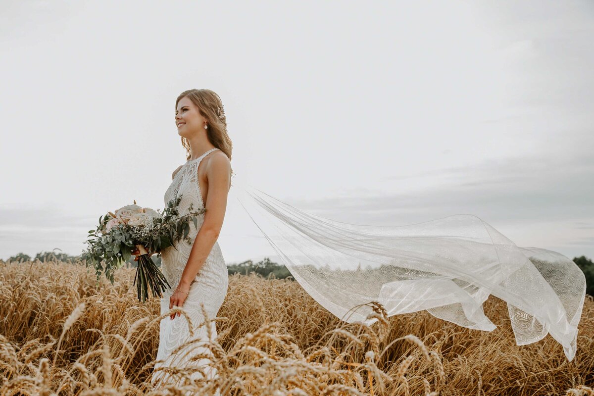 Bride standing in Exeter, Ontario field for wedding day photo. She is side profile to the camera looking off in the distance with her veil blowing behind her in the wind.
