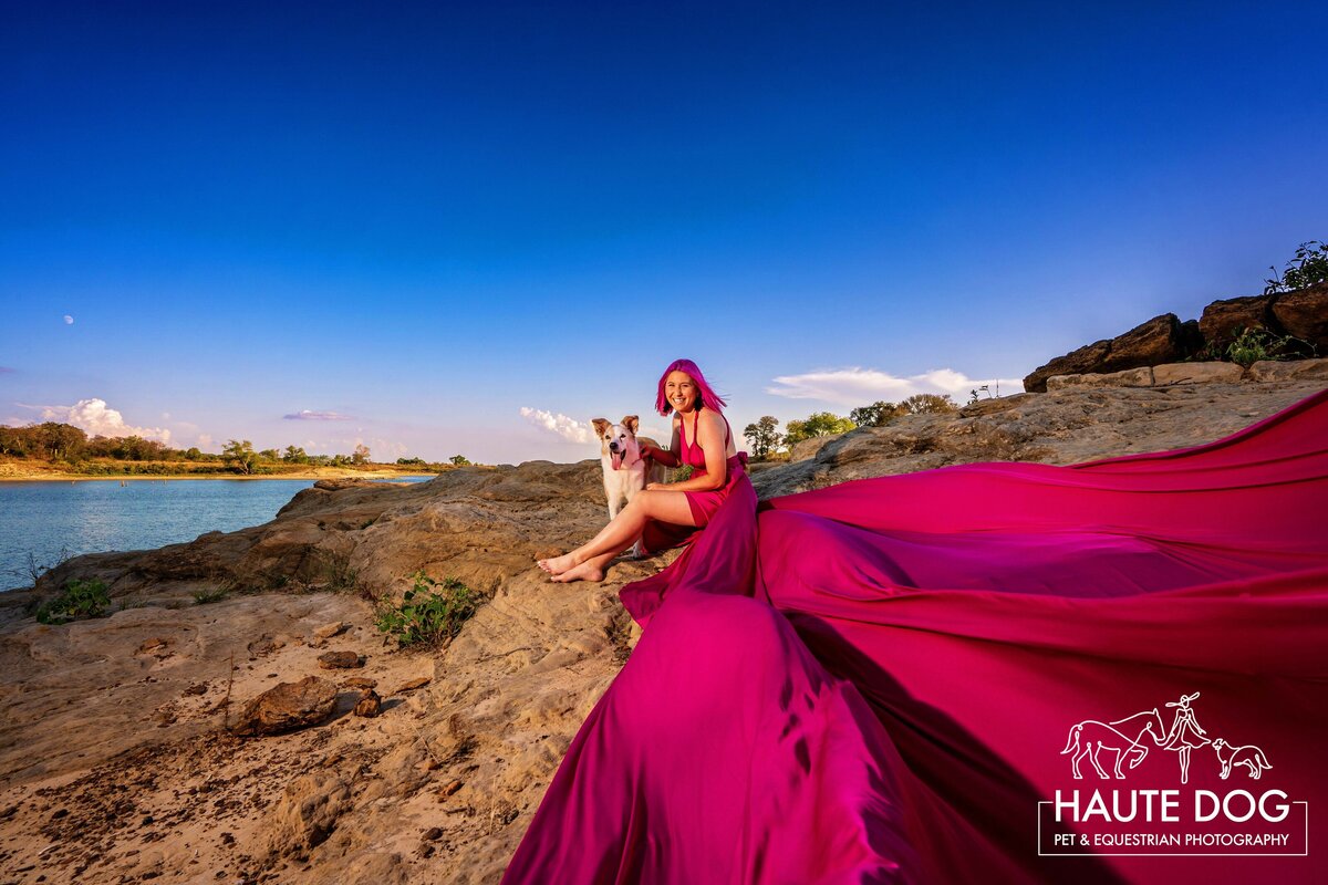 A woman with pink hair wearing a long pink flying dress sits on the edge of Grapevine Lake with her dog.