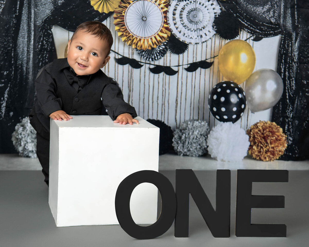 A baby smiles in a black shirt leaning on a white box for first birthday