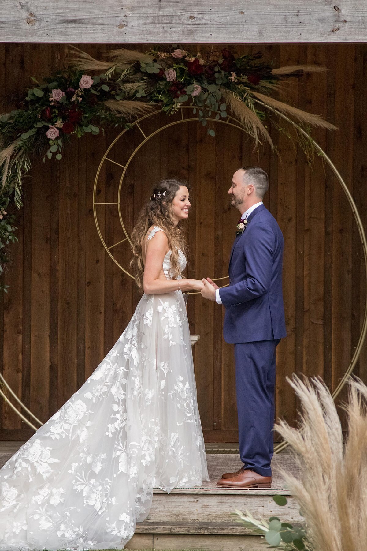 The bride, wearing a long sleeveless lace wedding dress with a plunging neckline holds hands with the groom wearing a navy suit and burgundy tie under the rustic log wedding pavilion at Saddle Woods Farm. They are standing in front of their gold circular ceremony arbor with a crescent moon and boho burgundy and blush wedding flowers with pampas grass.