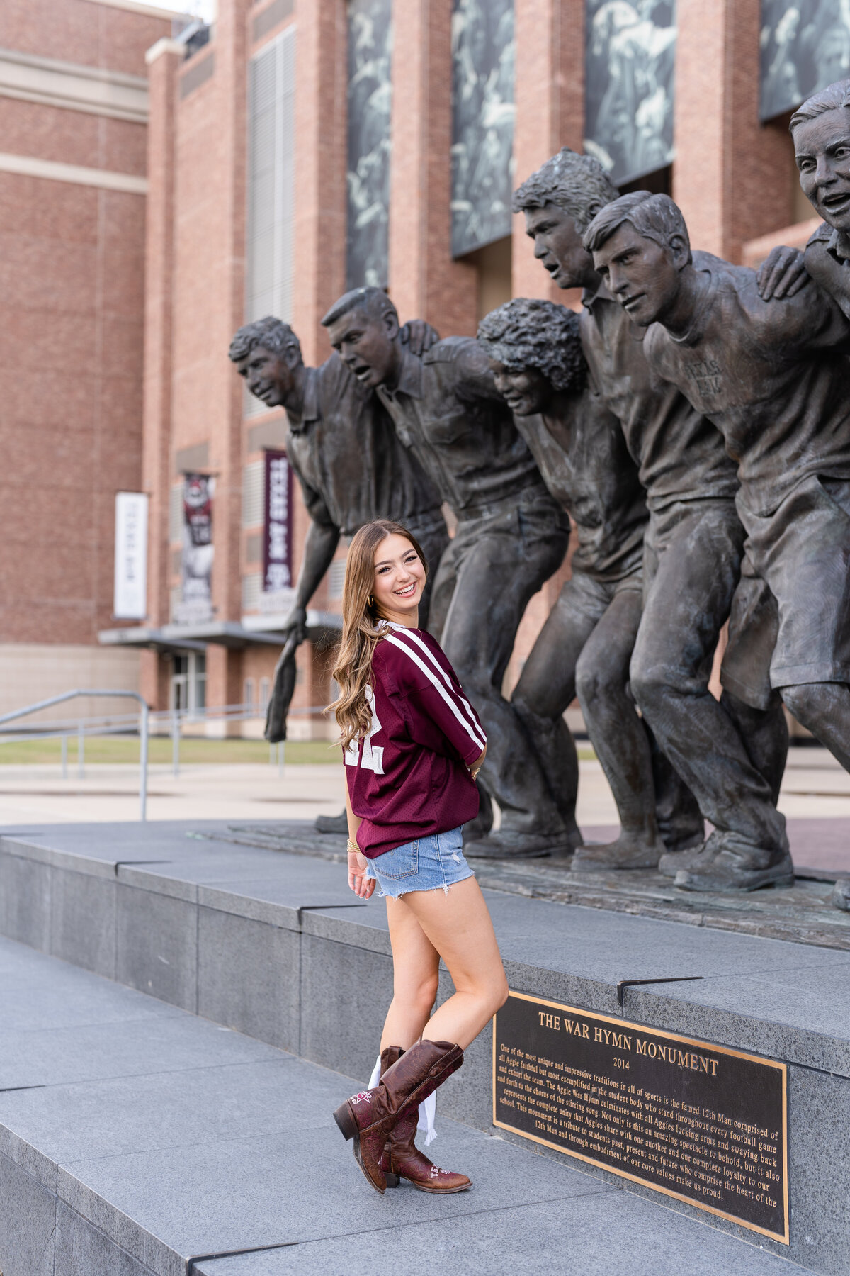 Texas A&M senior girl laughing and twirling around while standing in front of war hymn statue while wearing maroon jersey and cowboy boots at Kyle Field