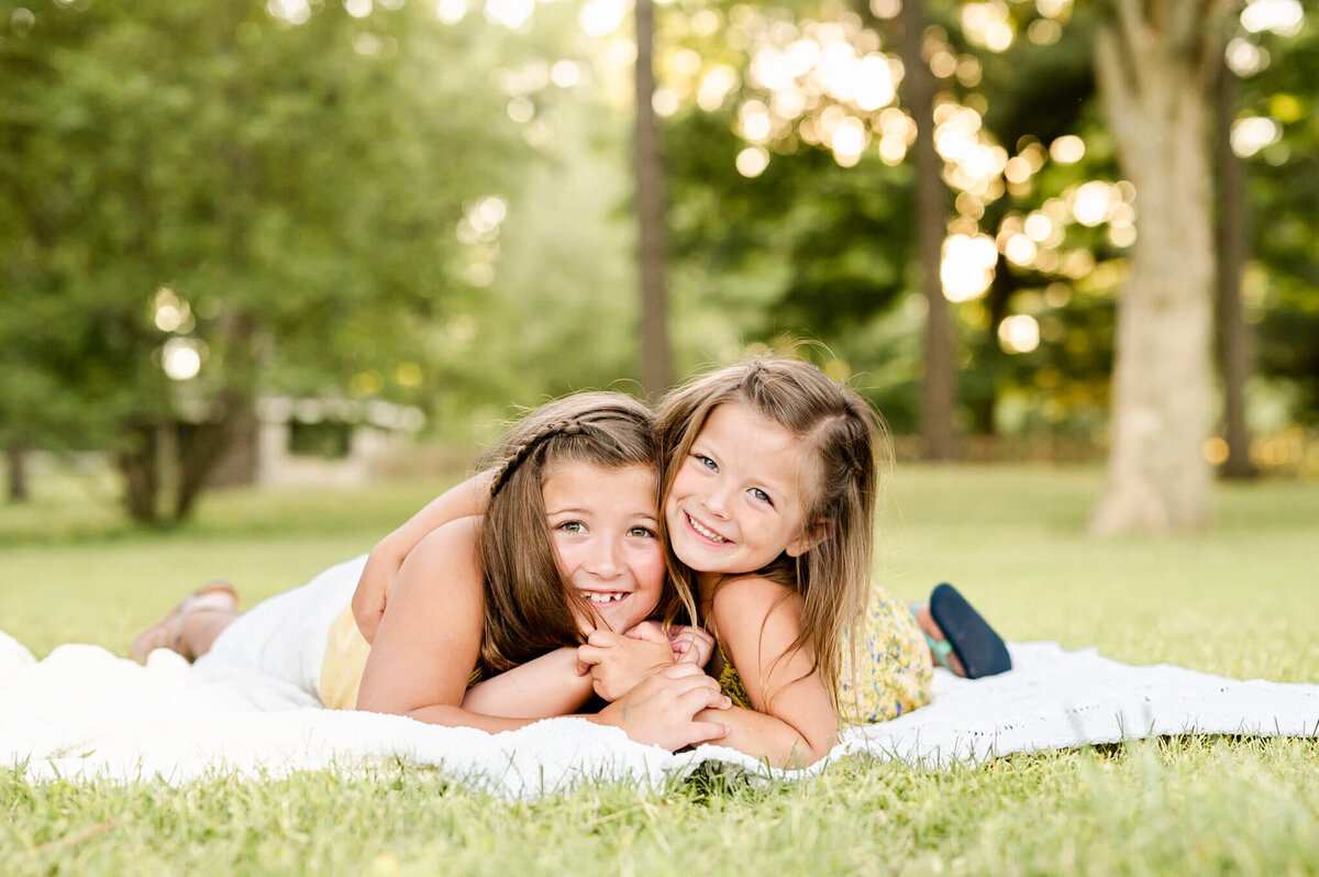 Silly sibling picture of two sisters hugging on a blanket in a park for family photos near Geneva.