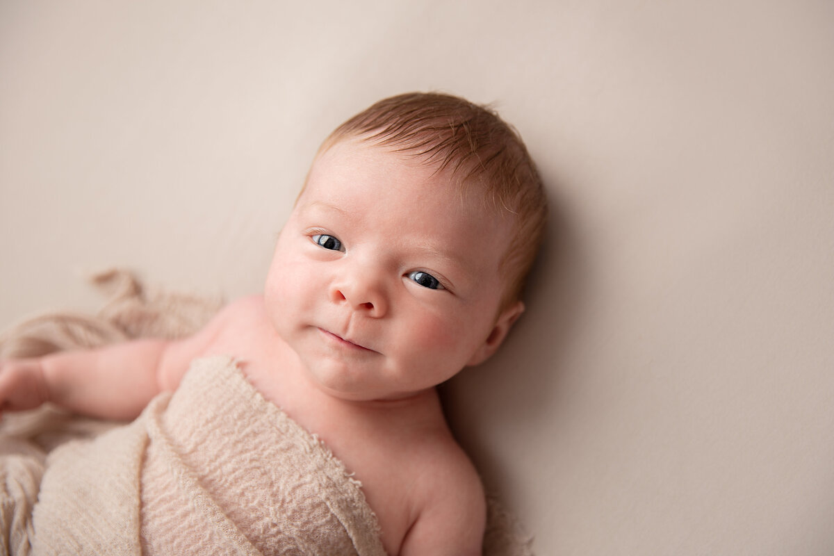 Baby lying on his back on a beige background with a beige wrap over him and looking straight into the camera.