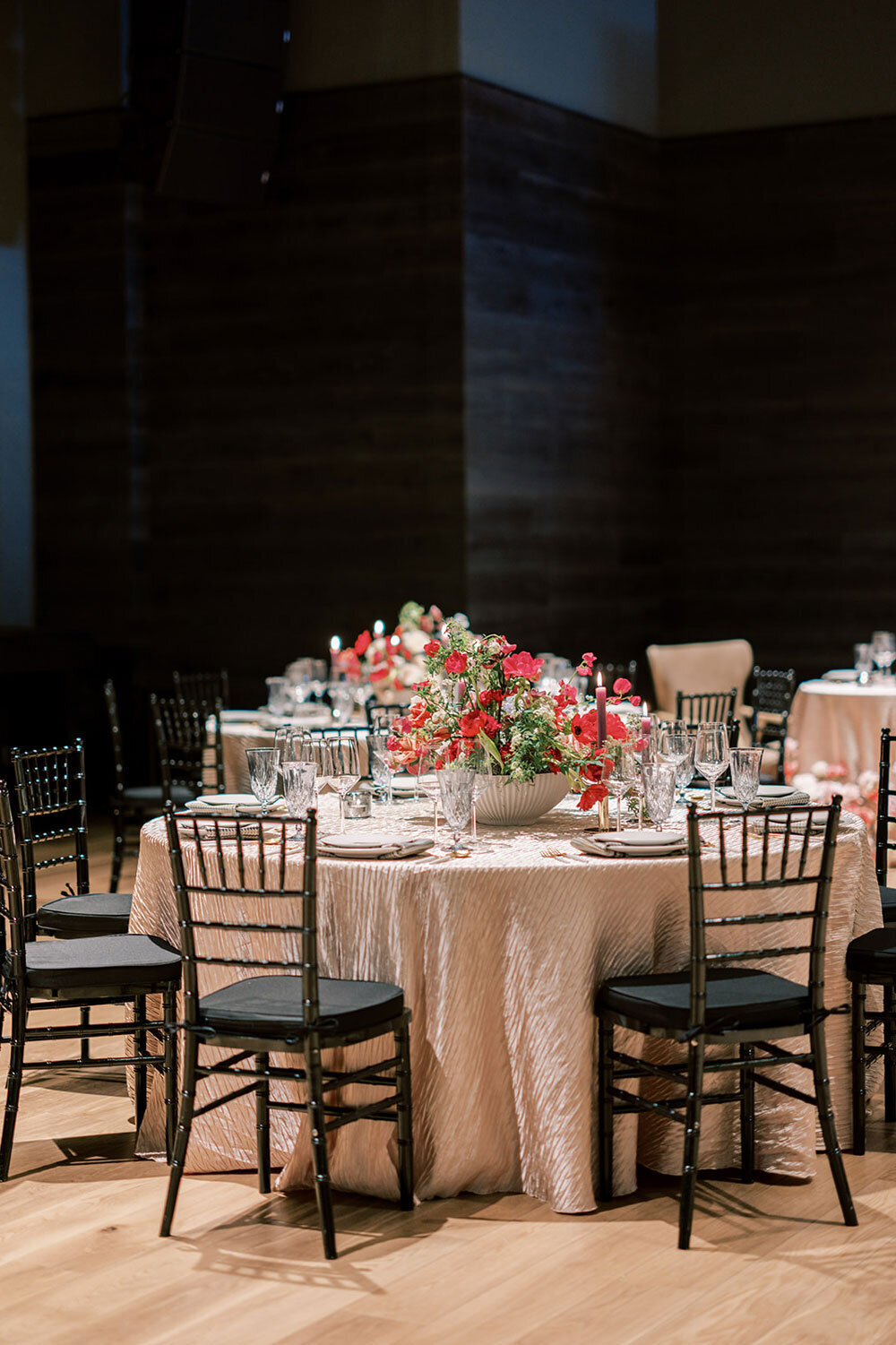 Round wedding reception table with blush textured linen, black chairs, and a centerpiece of vibrant red and pink florals.