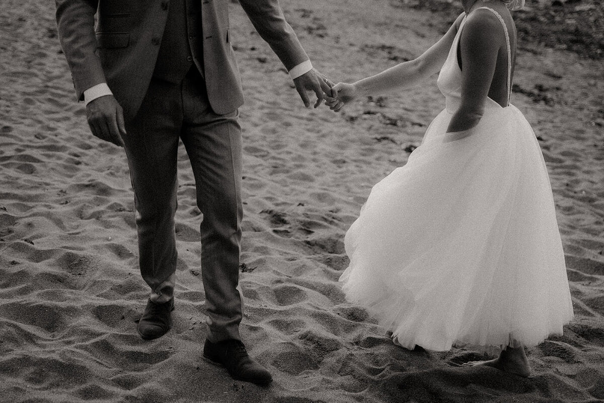 Mariés se tenant la main sur le sable, avançant ensemble. Photo en noir et blanc par Kamescop.