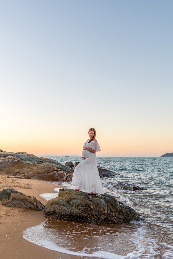 pregnant woman in white gown standing on a rock at the beach at sunset - Townsville Maternity Photography by Jamie Simmons