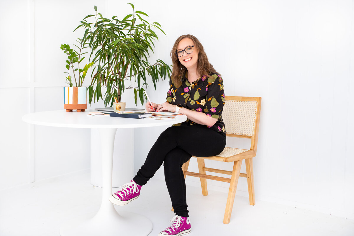Melissa-Packham_A-Brand-Is-Not-A-Logo_Melissa-sitting-at-table-on-cane-chair-plants-in-corner_wearing-floral-top-black-jeans-magenta-converse-sneakers_Landscape
