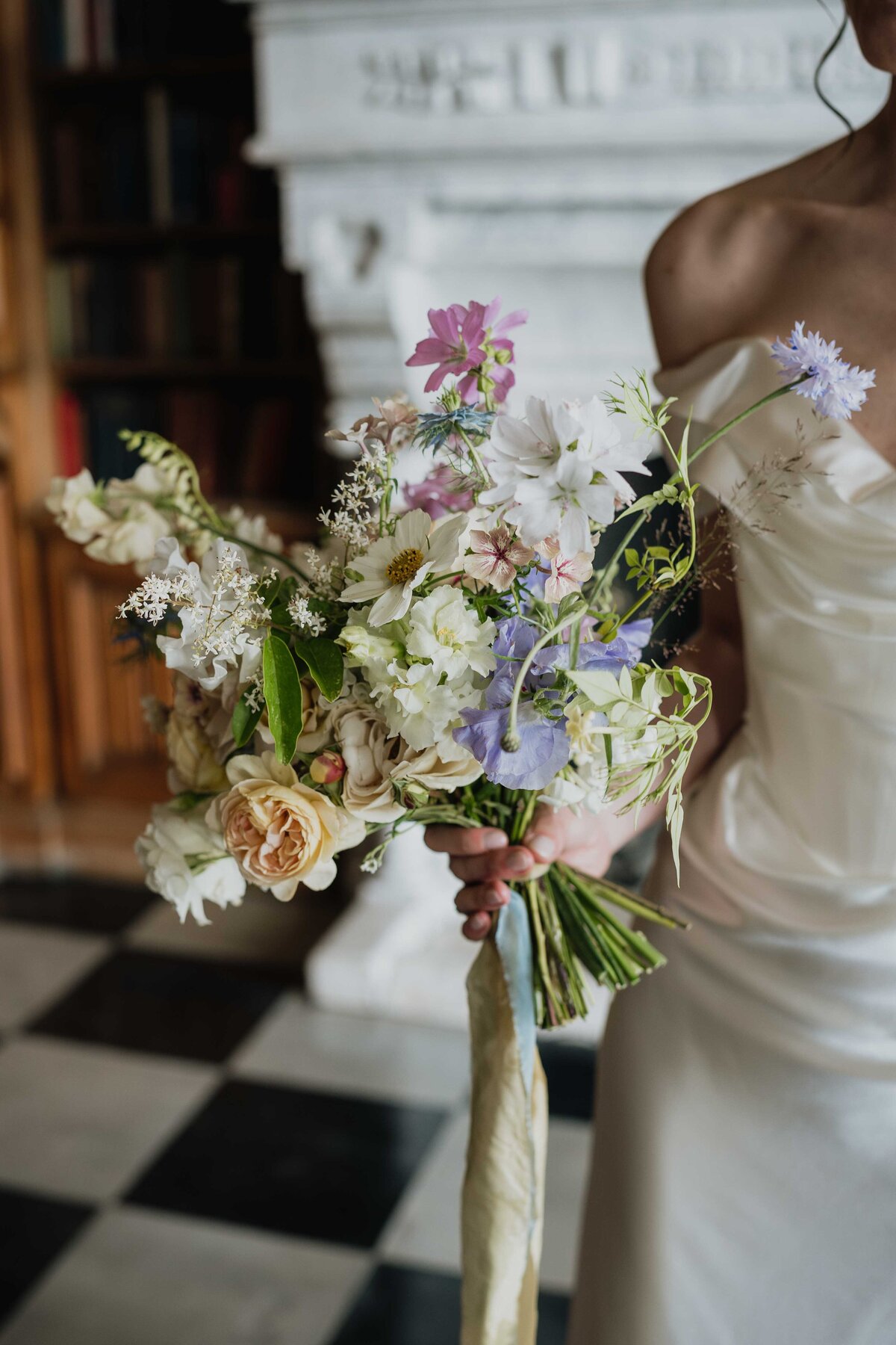 A bride in an elegant white Vivienne Westwood wedding gown holds a bouquet of assorted flowers, including roses and wildflowers. Photographed beautifully by her wedding photographer, the scene features a black and white checkered floor and a white fireplace adorned with bookshelves.