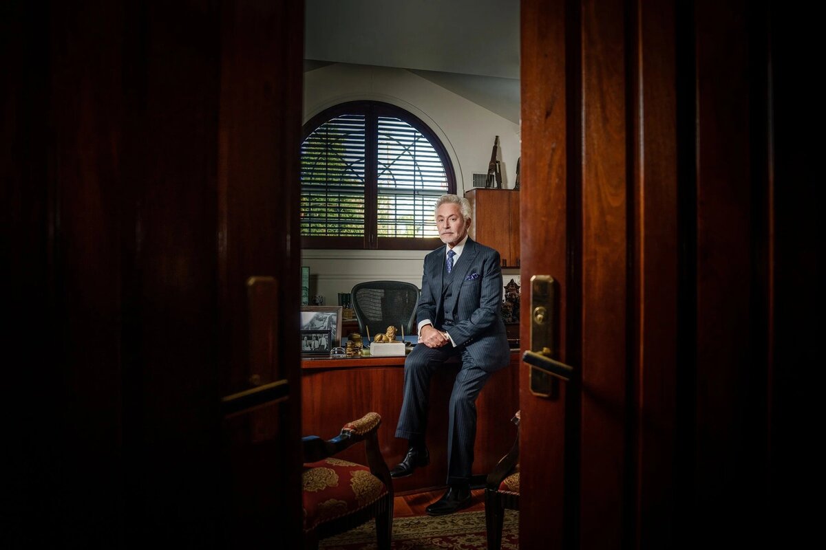 Professional headshot of businessman seated confidently at large wooden office desk with window background new york.