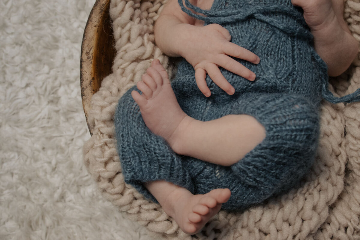 Close-up of a newborn's tiny feet peeking out from a blue knit romper, lying on a fluffy white surface