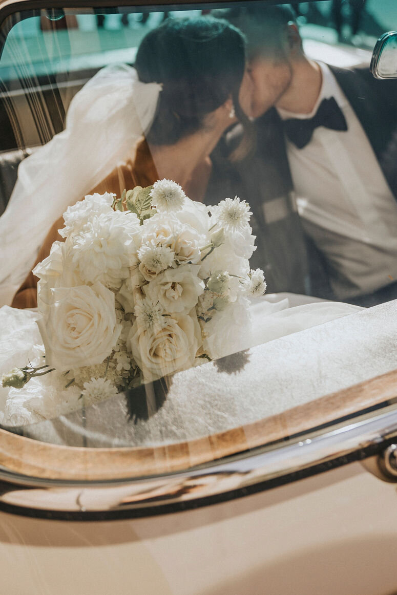 A bride and groom kiss in a limousine with a white bridal bouquet in the foreground.