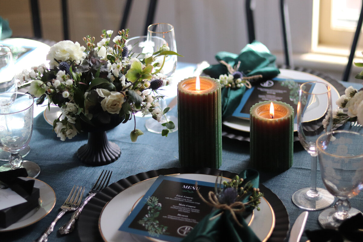 A black blue green and white wedding reception table with preset menus, pillar candles, and green velvet napkins