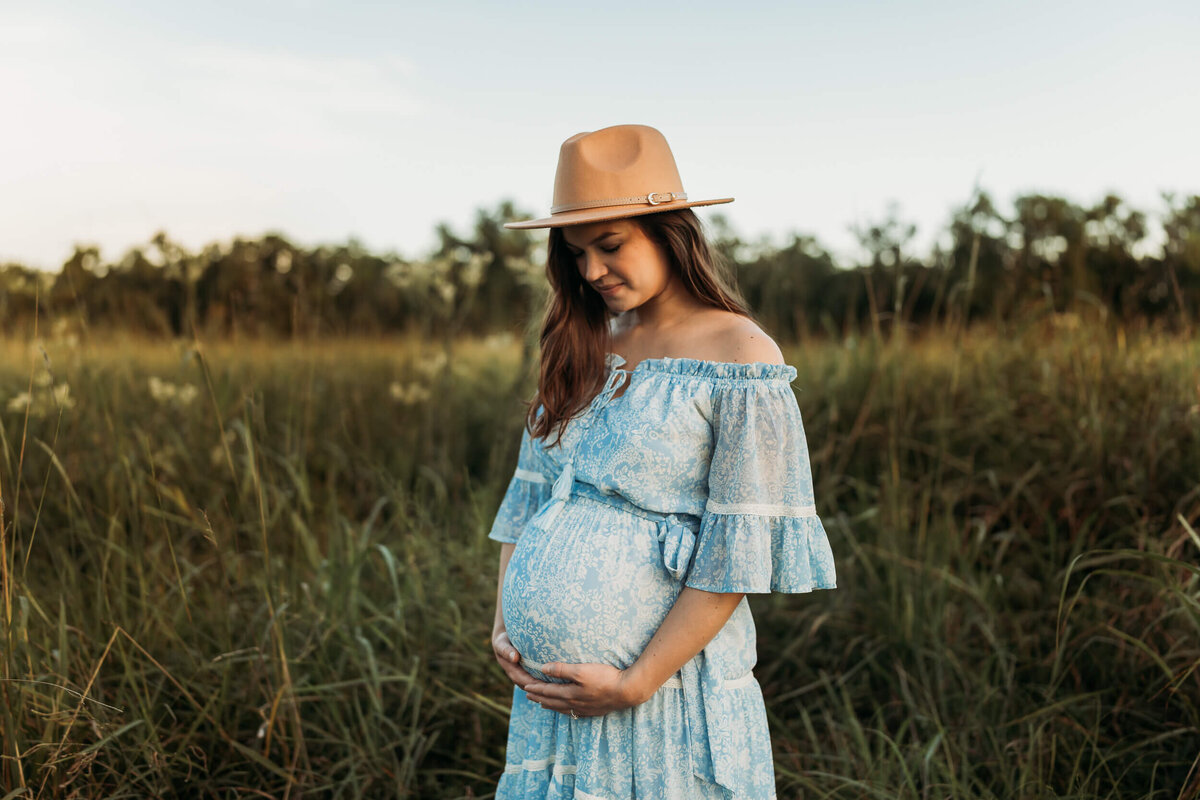 gorgous expectant mama holds her belly in a field, wearing a hat.