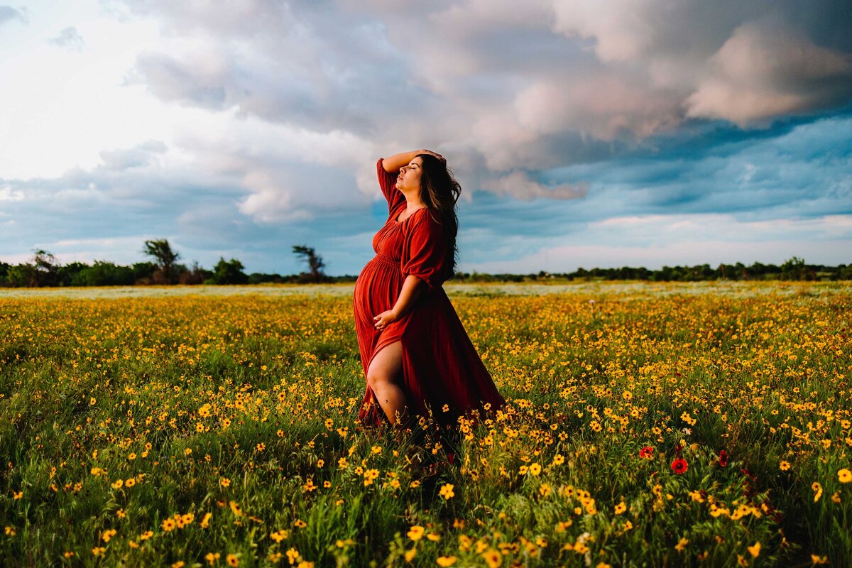 The future mom, dressed in a red long dress, joyfully poses in a beautiful garden adorned with yellow flowers. She radiates happiness as she prepares for her new baby.