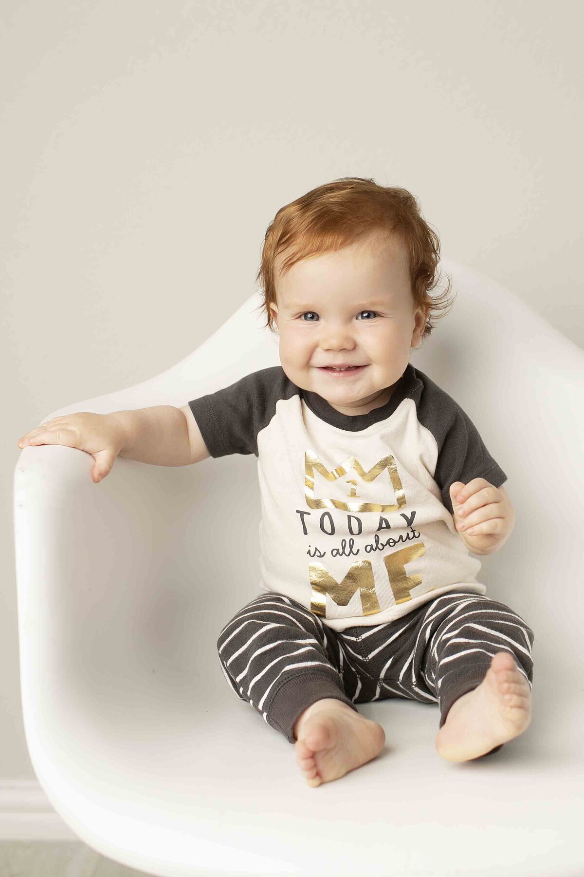 Birthday boy with red hair sitting in a white chair, dressed for his special day in a simple studio backdrop