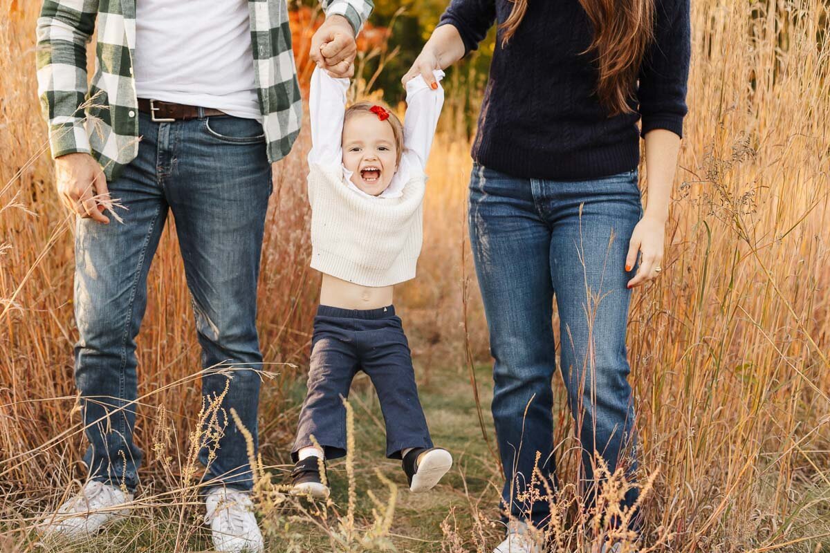 A child in a white sweater and blue pants is joyfully swinging between two adults holding hands in a grassy field, captured beautifully by a Toronto lifestyle family photographer.