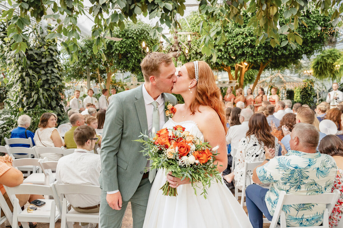 The bride and groom share a kiss at the end of the ceremony, surrounded by wedding guests in the lush greenhouse venue of Le Jardin.