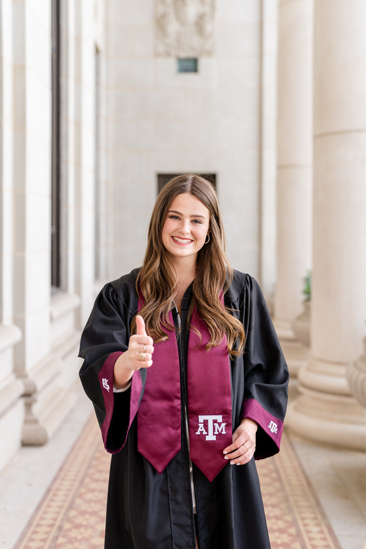 Texas A&M senior girl wearing gown and Aggie stole while holding thumbs up and smiling in middle of columns of Administration Building