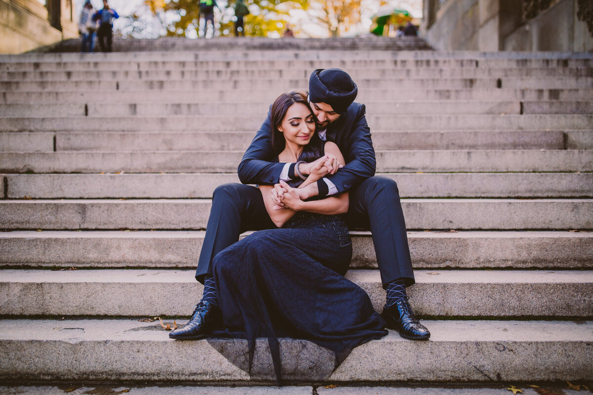 A couple sitting on the stairs embracing during their engagement session in all black clothing at central park bethesda terrace