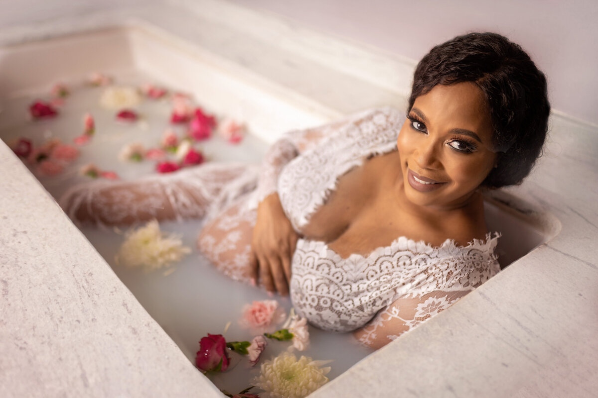 Pregnancy milk bath with purple, white,  and red flowers.   A smiling African American woman is half submerged in a milk bath tub, holding her belly and smiling at the camera.  Her dark hair is pulled up.  She is wearing a white lace dress.