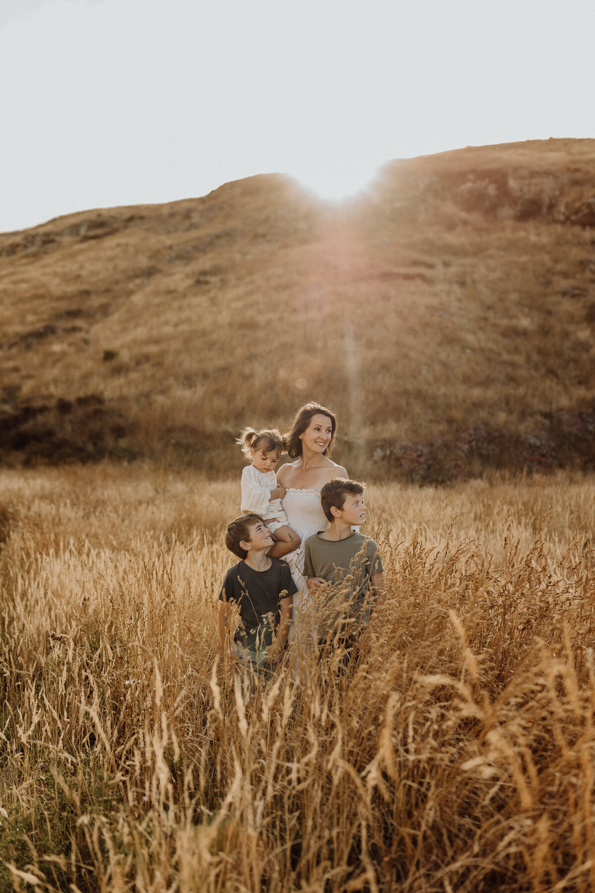 A mother and her three children standing in waist high grass while the sun shines in the background during a family photoshoot in christchurch