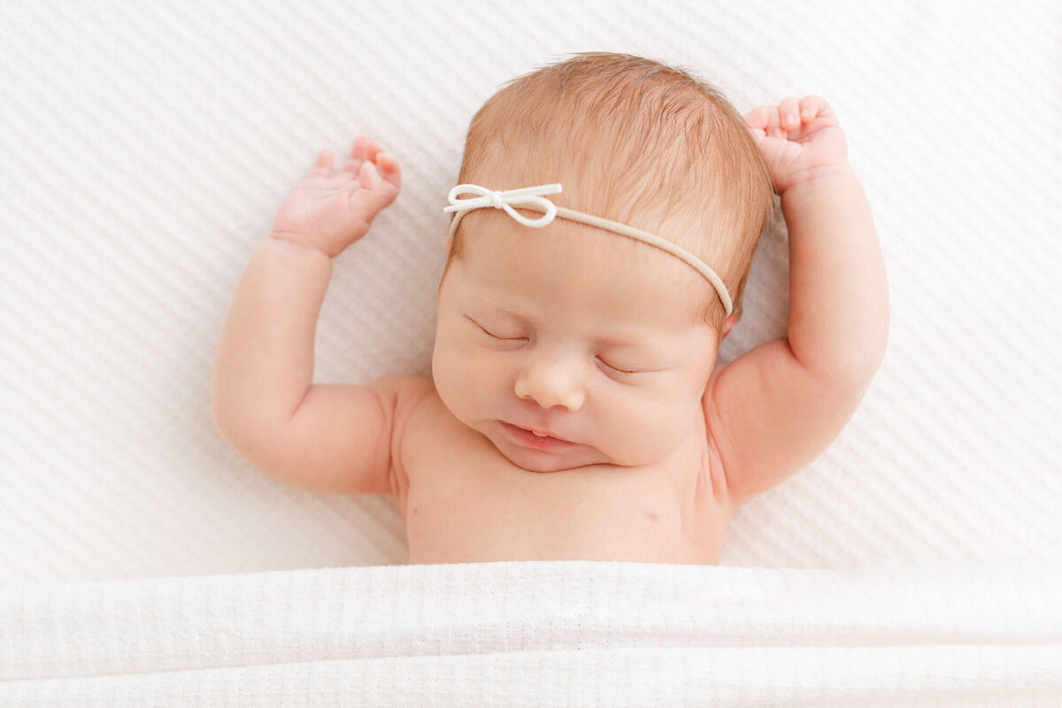 Baby sleeping on textured white blanket and covered up to her upper belly by blanket. She is sleeping and has her hands up by her head. She is wearing a minimalist white bow. Image by Ashlie Behm Photography