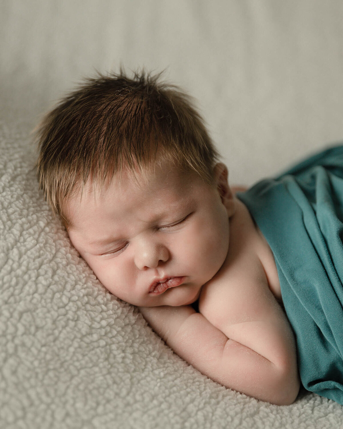 Newborn boy with lots of light brown hair laying on his stomach with a teal wrap and creme background.