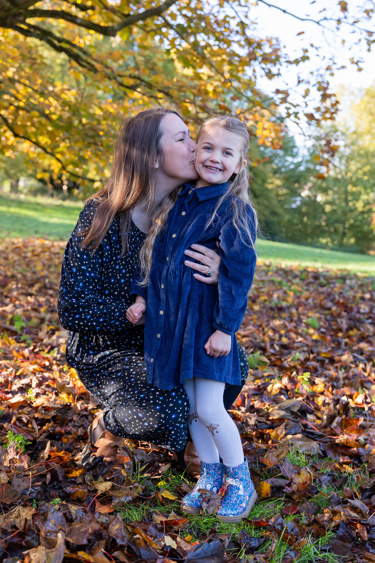 A woman kissing a young girl on the cheek in a park filled with autumn leaves. both are smiling and dressed in blue.