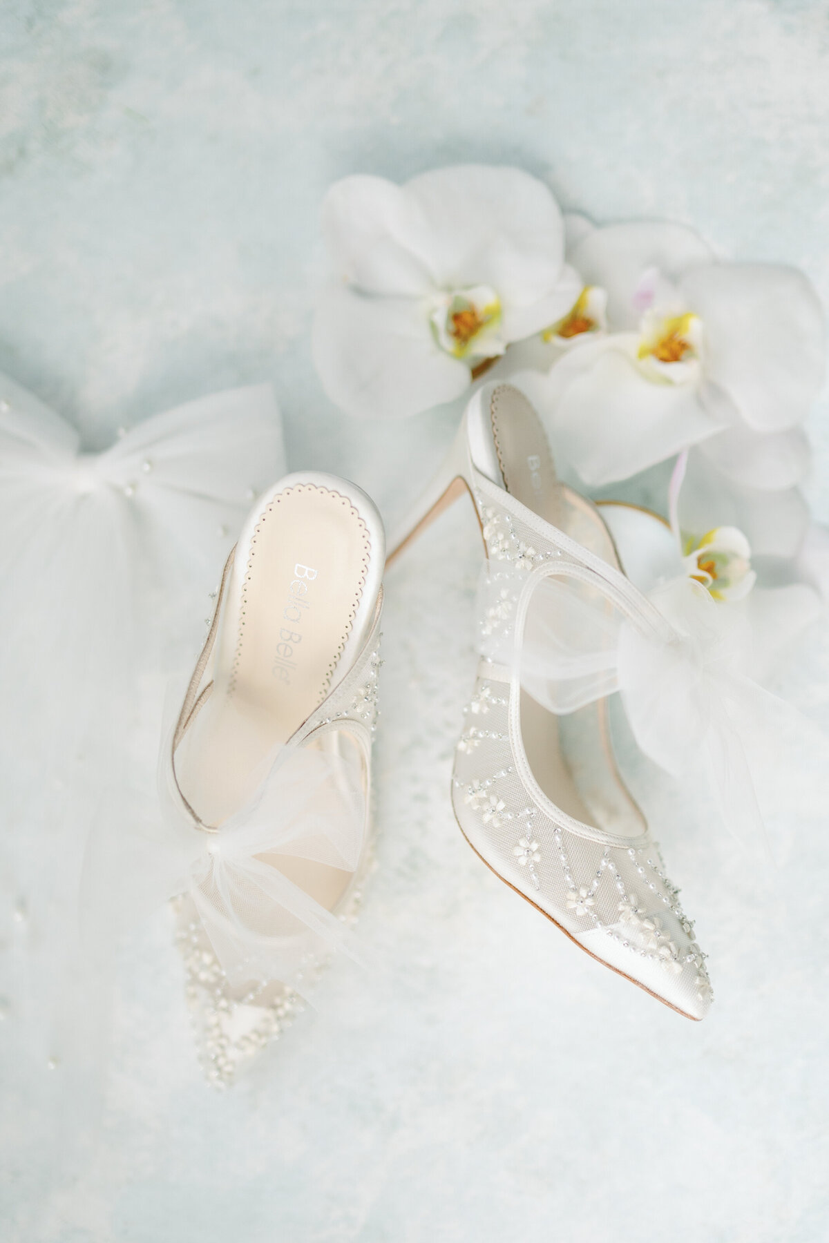 A classic wedding detail shot featuring elegant white bridal shoes paired with stunning white orchids. This sophisticated combination symbolizes purity and grace, perfect for a timeless and luxurious wedding aesthetic.