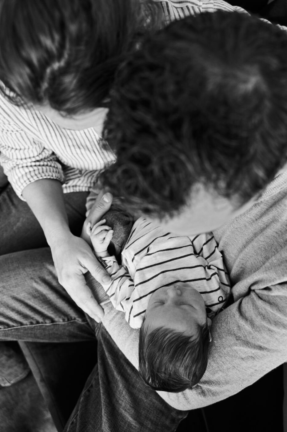  A black and white, high-angle photo of a newborn baby being held by a parent. The baby is wearing a striped outfit and is held gently in the parent's arms. The parent's hair and clothes are visible in the foreground, creating a loving, intimate composition.