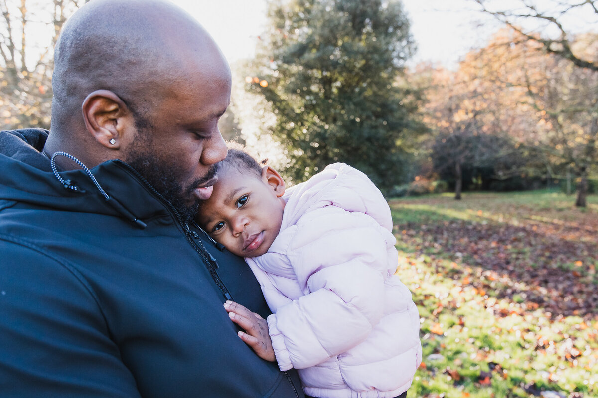 A man tenderly holding a young child in a park on a sunny day. both are partially facing the camera, with trees in the background lit by sunlight.