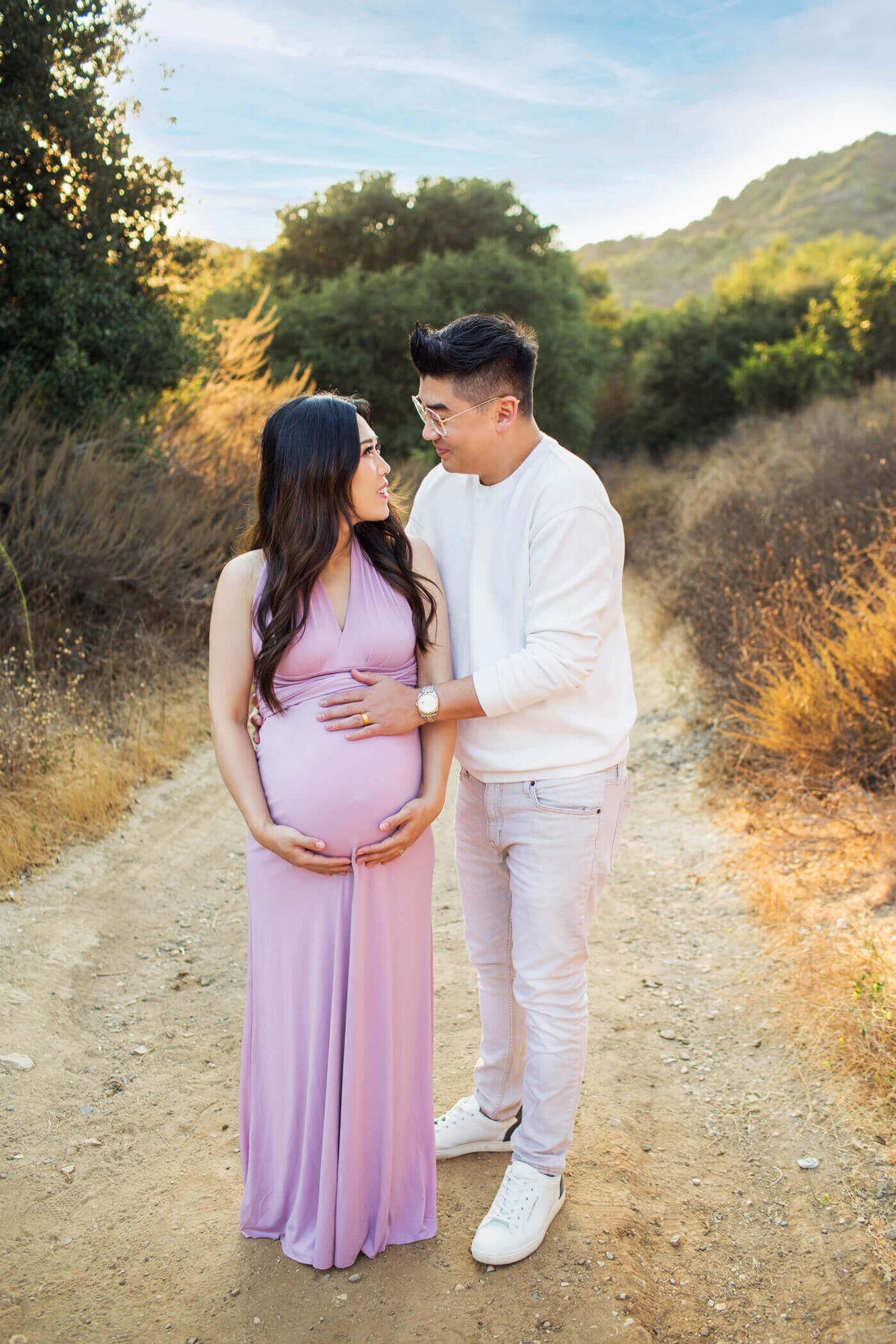 A pregnant woman stands on a dirt path, lovingly embraced by her partner. She is wearing a form-fitting, sleeveless lavender dress that accentuates her baby bump, while he is dressed in a white long-sleeve shirt and light pants. They gaze into each other's eyes, sharing a tender moment. The surrounding greenery and golden sunlight create a warm and serene atmosphere.