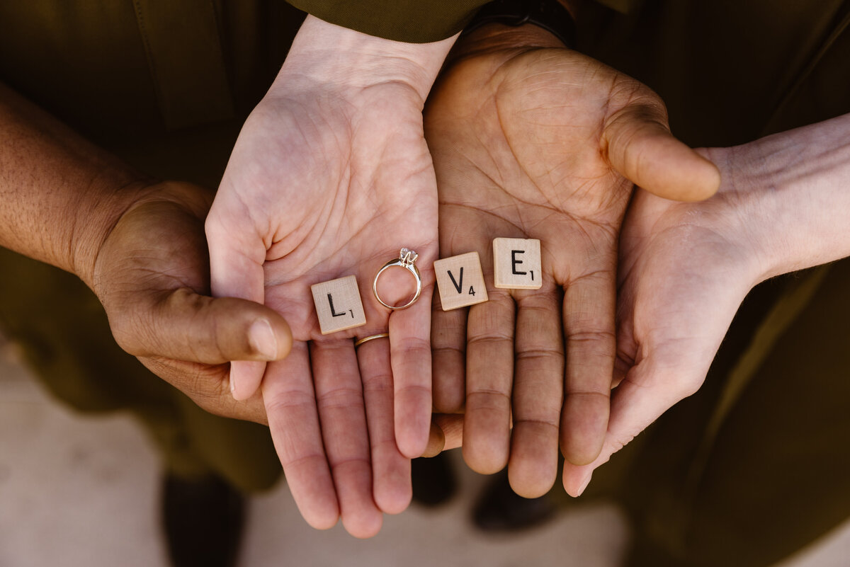 A couple’s hands hold Scrabble tiles spelling “LOVE” with an engagement ring replacing the letter O, highlighting their engagement in a meaningful and creative way.