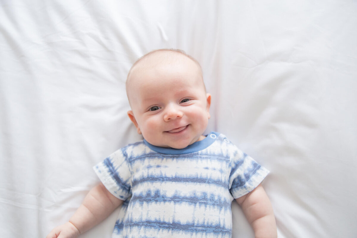 view from above of Happy infant baby boy in blue and white tye dye shirt  on a white bed during session with las vegas milestone photographer