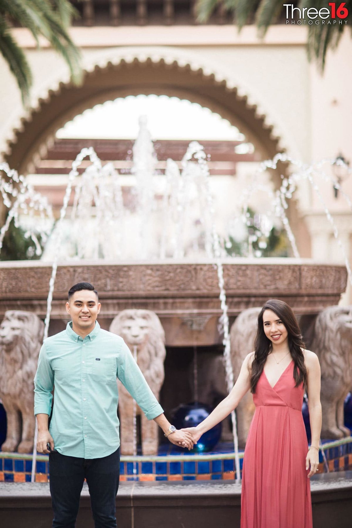 Engaged couple hold hands as they pose in front of water fountain