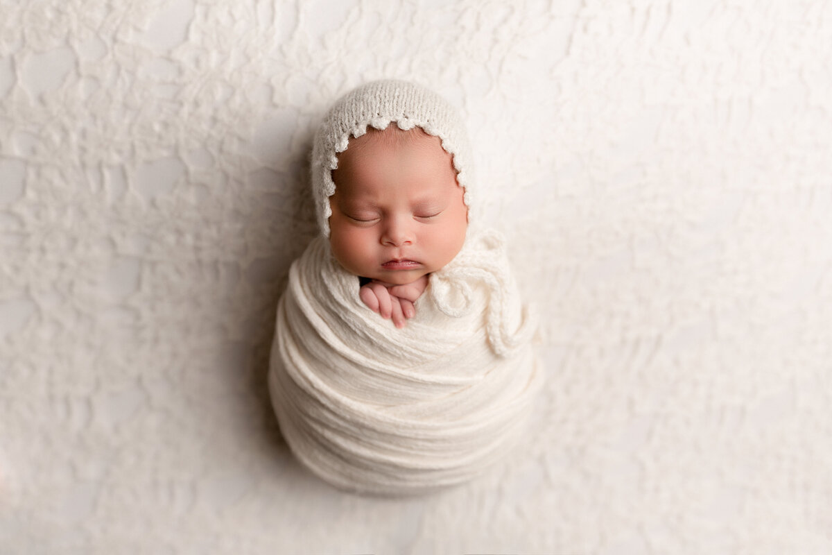Newborn photos from top West Palm Beach newborn photographer - baby is swaddled in a cream knit wrap with a matching crocheted bonnet. Baby's fingers are peeking out of the top of the wrap. Baby is sleeping.  Aerial image.