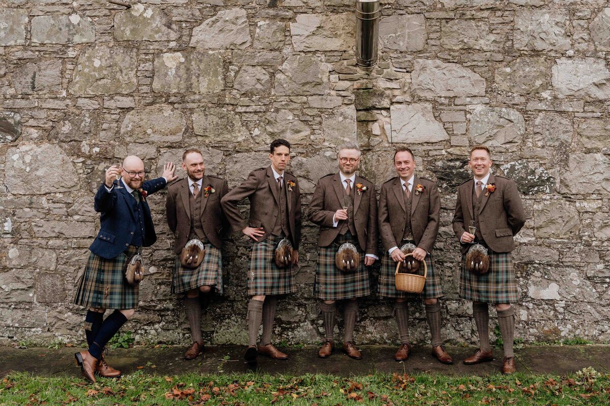 Six men in kilts and tweed jackets pose cheerfully against a stone wall at Charleton Fruit Farm, Aberdeen. Dressed in traditional Scottish attire, some hold drinks while one holds a basket. The scene is outdoors with fallen leaves scattered on the ground, they smile towards a wedding photographer.