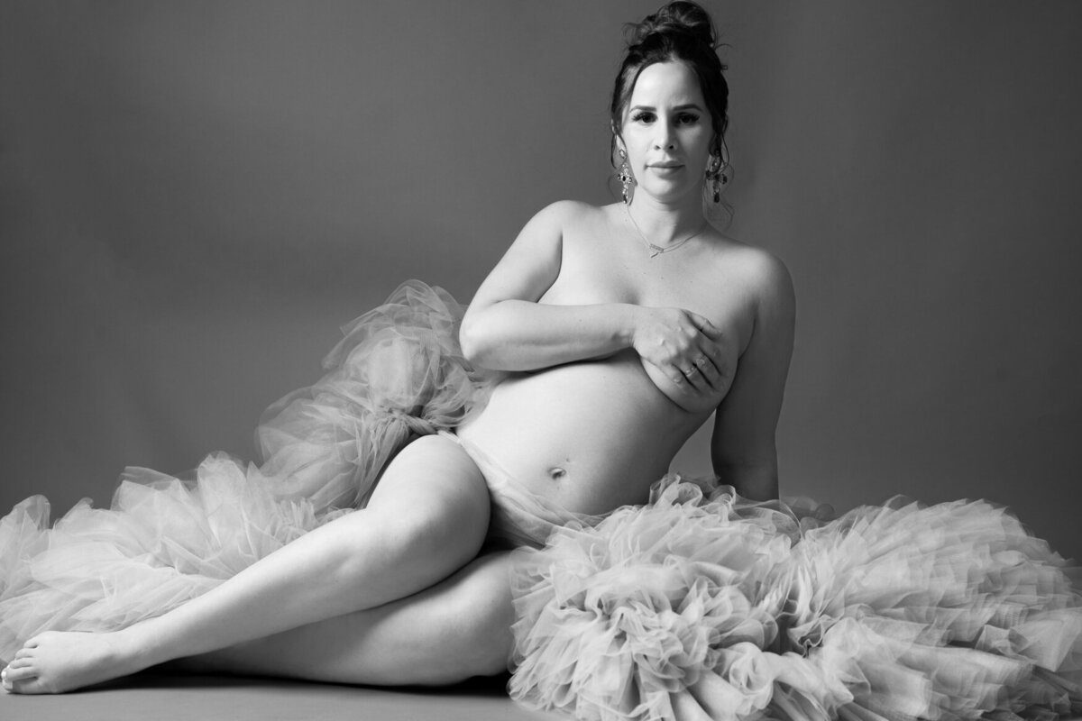 A black-and-white maternity portrait featuring a pregnant individual reclining gracefully against a backdrop of voluminous tulle fabric. The subject sits with legs extended and crossed, one hand resting on their baby bump while the other covers their chest.